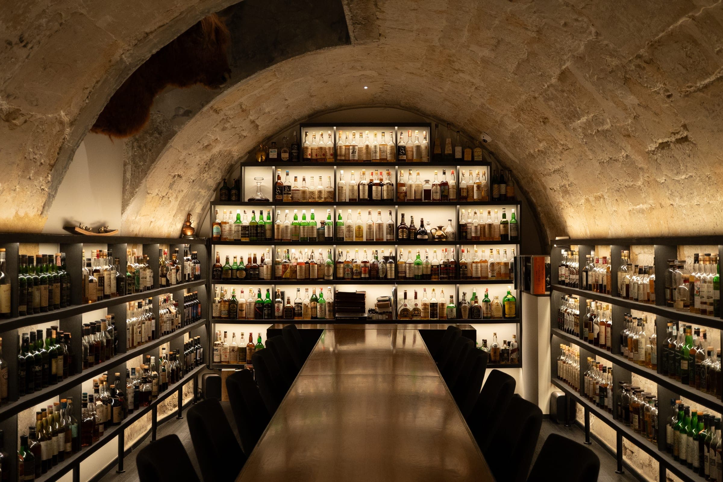Cave à whisky voûtée du bar Golden Promise, étagères illuminées et longue table en bois