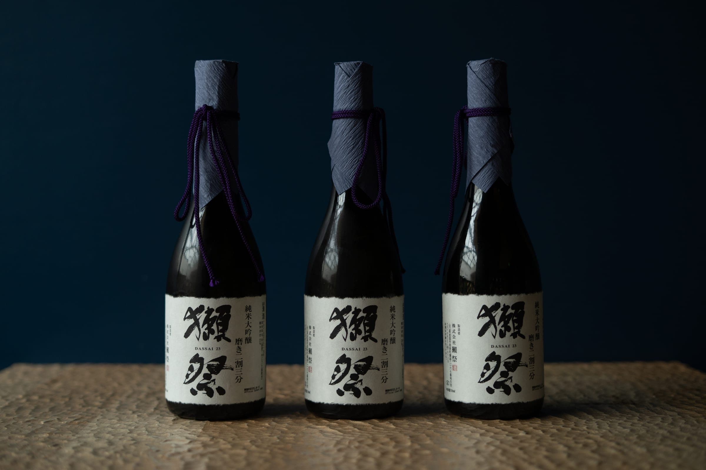 Trois bouteilles de saké Dassai, cols noués de tissu bleu indigo, sur fond sombre